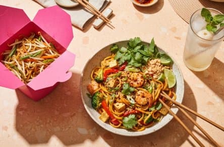 Noodles in a takeaway box next to a bowl of pad thai